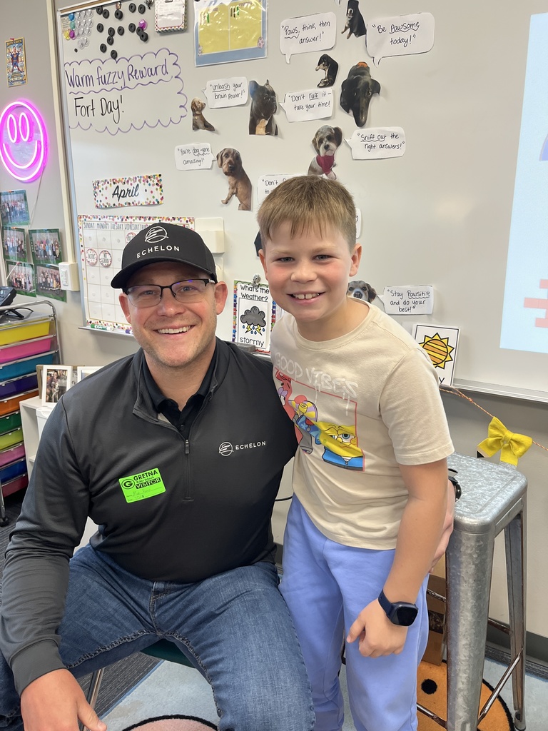 A man wearing a black "Echelon" hat and a visitor badge smiles while kneeling next to a young boy in a Spongebob Squarepants t-shirt in a classroom.