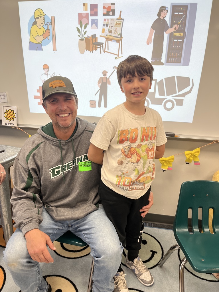 A man in a "Gretna" hoodie and a trucker hat smiles with his arm around a boy wearing a "Bo Nix" football t-shirt in front of a classroom projector screen.