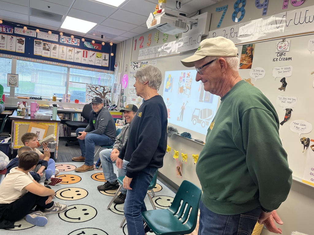 A group of adult visitors stand in a classroom, looking toward a group of elementary students sitting on a colorful "smiley face" rug.