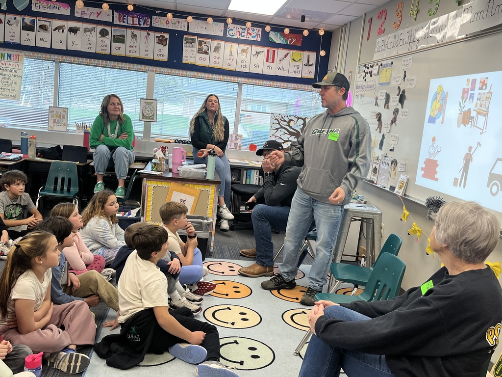 A man in a grey hoodie stands and speaks to a group of children sitting on a rug in a decorated classroom, while other adults listen.