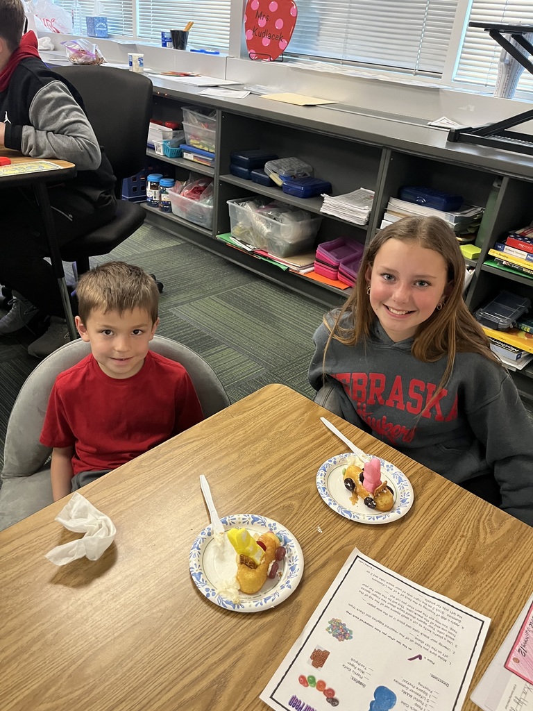 Two students sit at a wooden table with their completed candy-decorated snack cake cars and an instruction sheet.