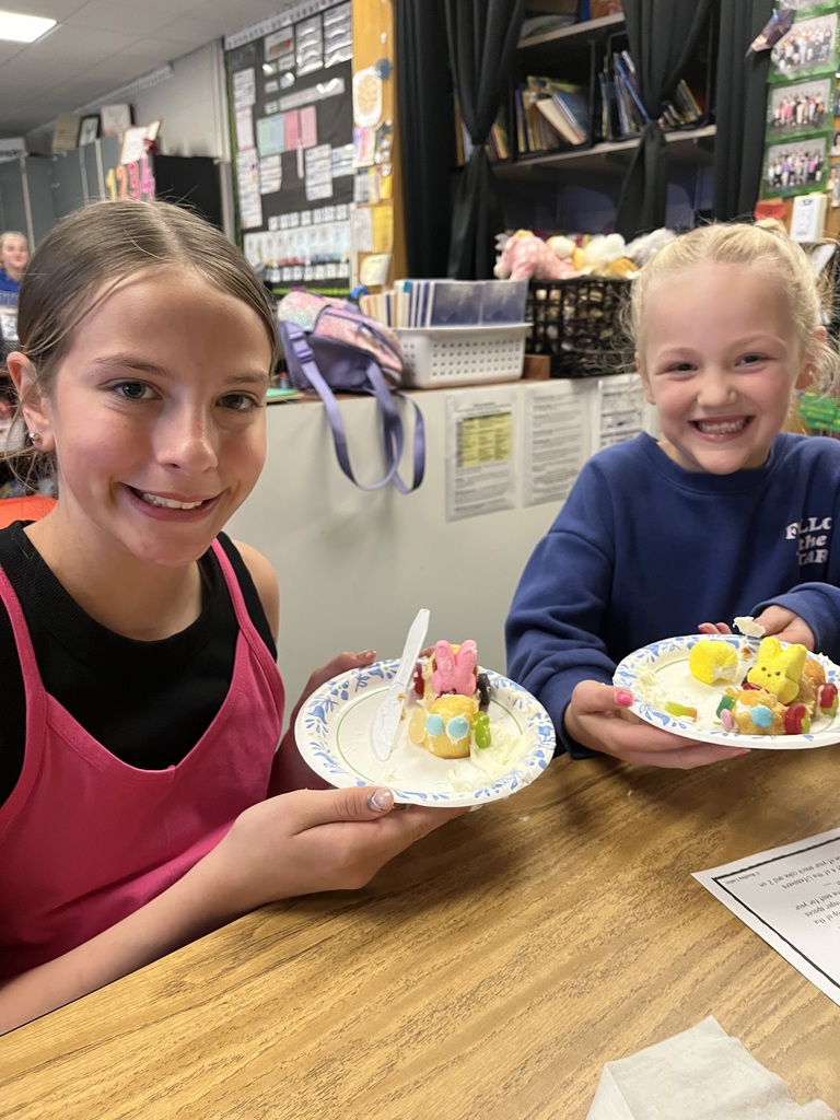 Two smiling girls at a desk show off their edible race cars made from snack cakes, frosting, and Peeps.