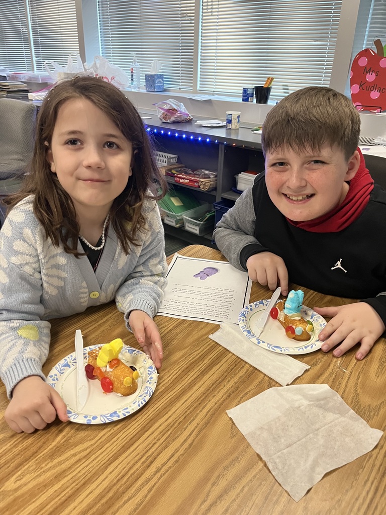 A girl in a floral cardigan and a boy in a black hoodie smile behind their Peep "race cars" in a classroom.