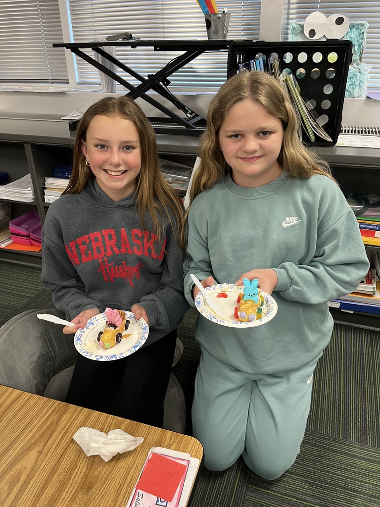 Two girls kneel on a classroom rug, proudly displaying their finished edible snack cake cars on paper plates.