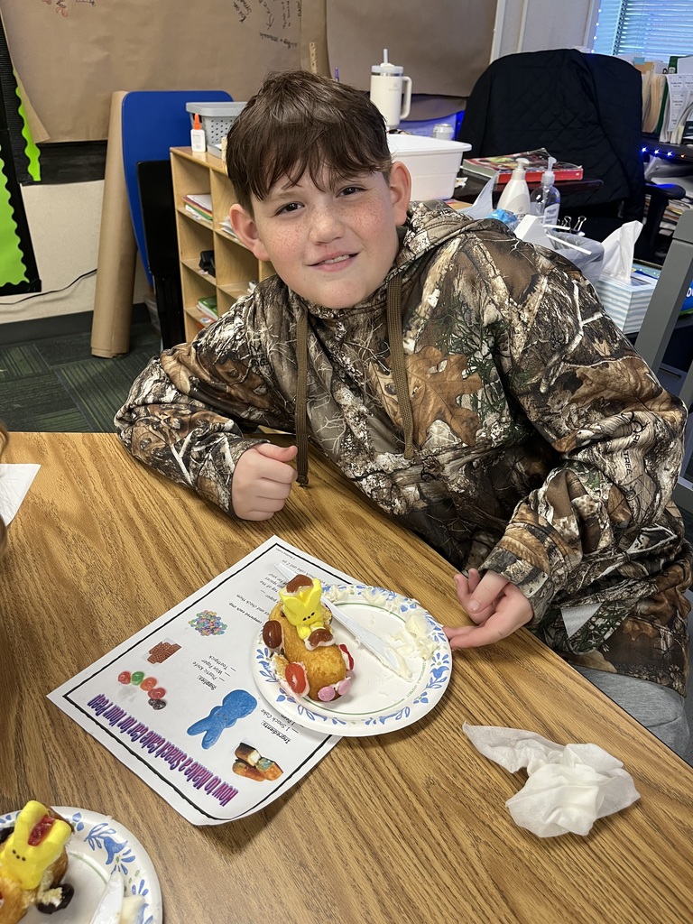 A boy in a camouflage hoodie sits at a desk next to his yellow Peep snack cake car and a project instruction sheet.