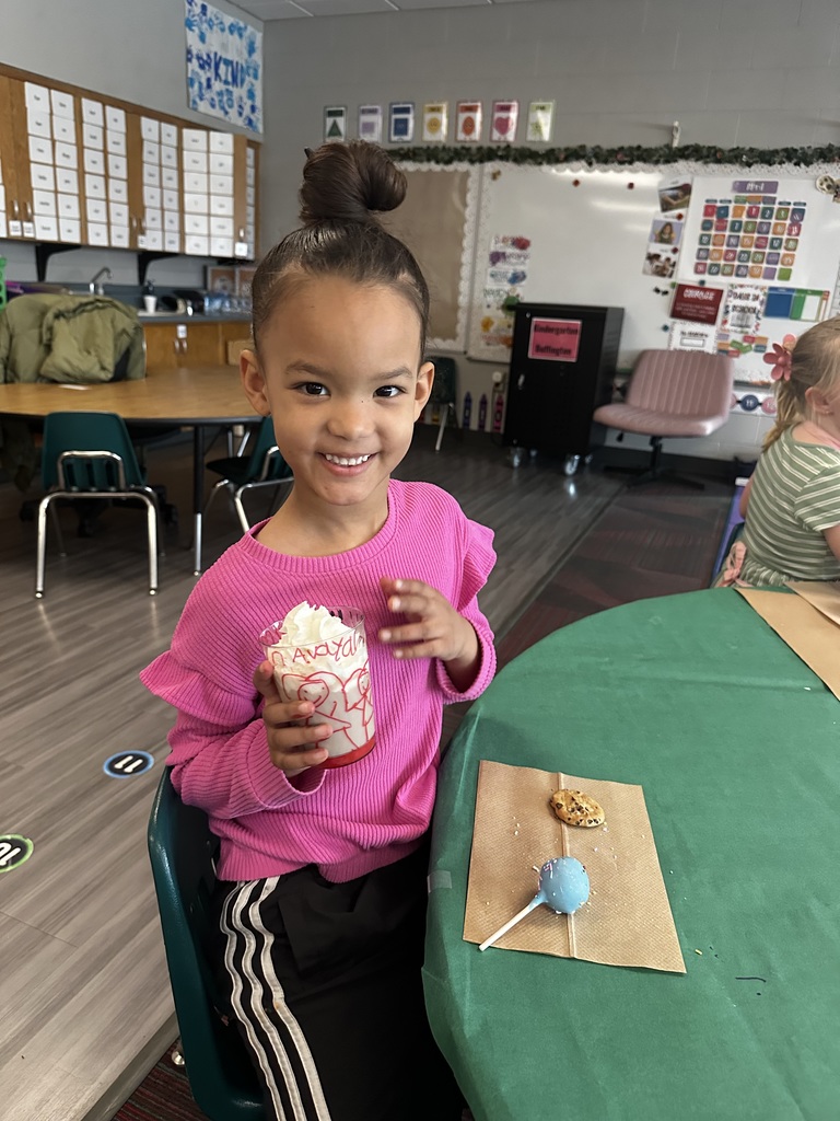 A young girl in a pink shirt smiles while holding a cup filled with whipped cream and a strawberry, next to a cake pop and cookie.