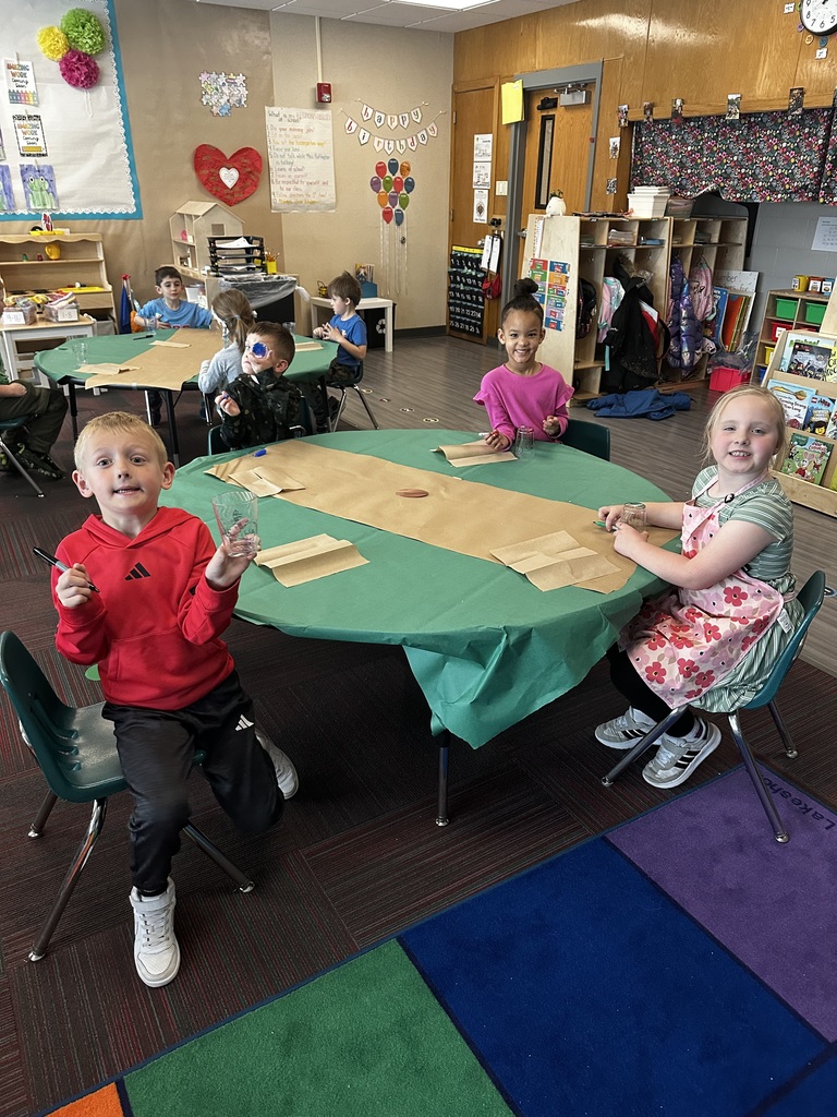 Three students sit at a table in a classroom with "Happy Birthday" decorations; one boy holds up his decorated cup.