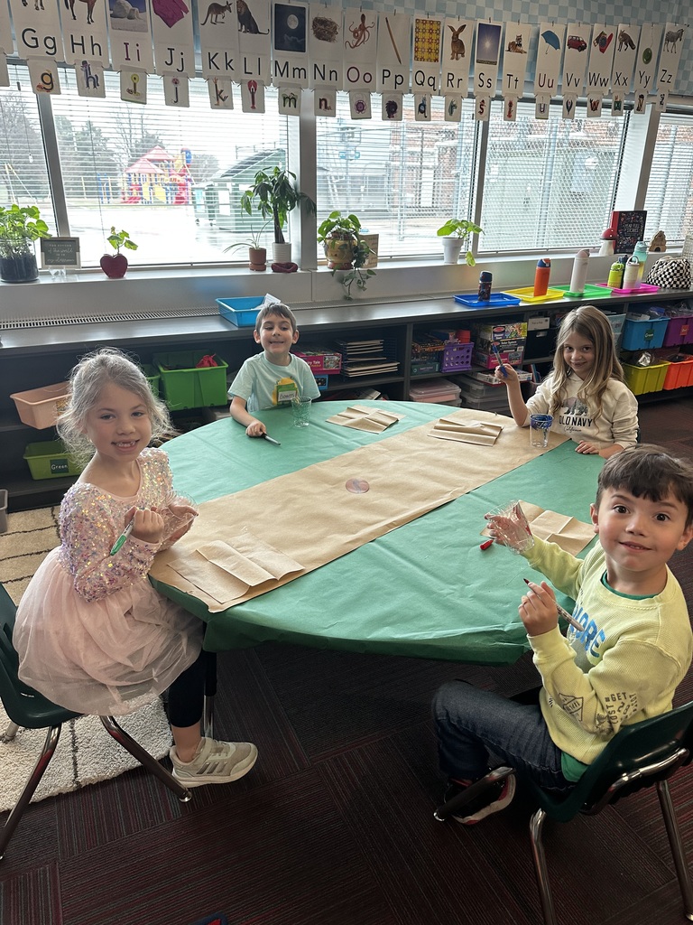 Four kindergarten students sit around a green circular table, using markers to decorate clear plastic cups.