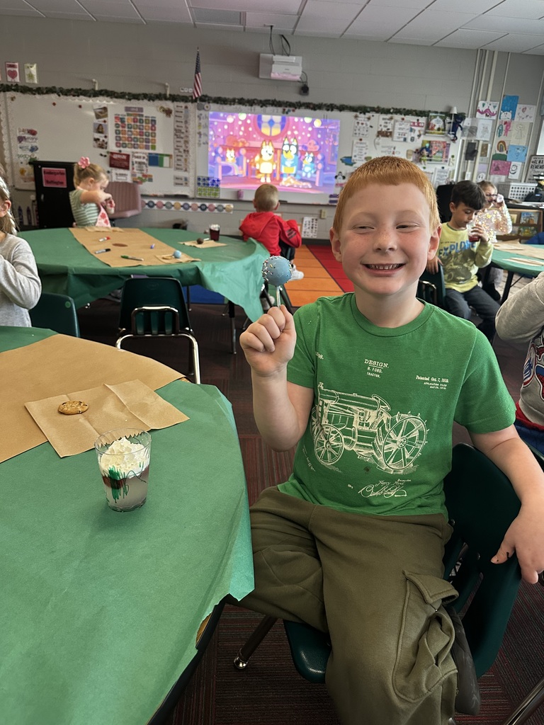A boy in a green shirt smiles broadly while holding a blue cake pop; a Bluey cartoon plays on a screen in the background.