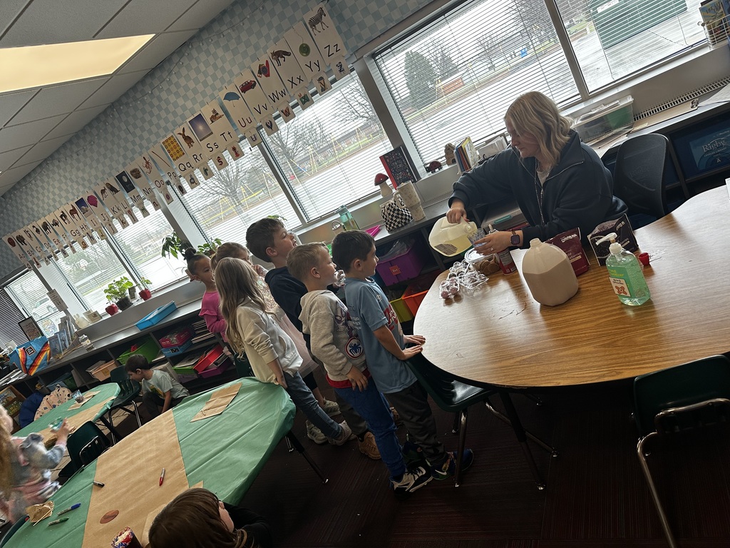 Students line up at a wooden table where a teacher pours milk into their decorated cups for a classroom treat.