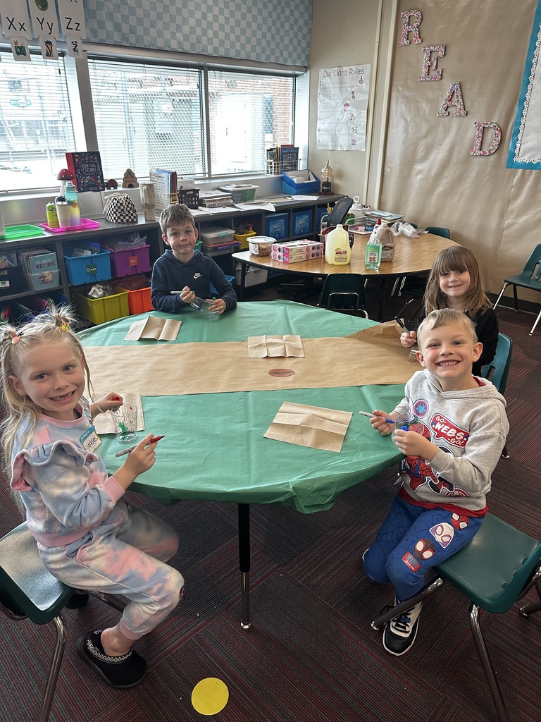 Four students smile at a green table while working on their cup-decorating project in a sunny classroom.