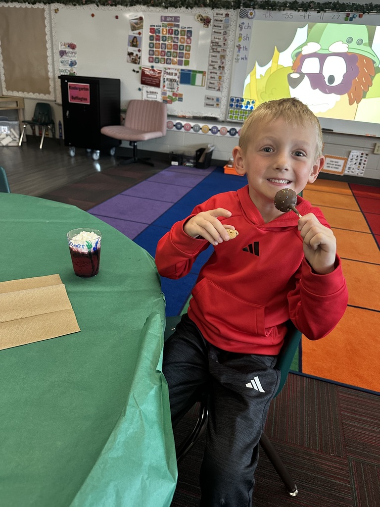 A boy in a red hoodie smiles while holding a chocolate cake pop, with his decorated cup sitting on the table.
