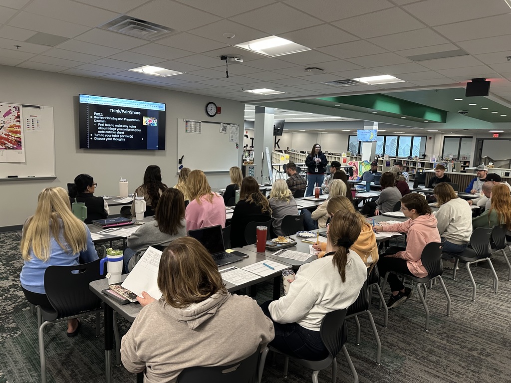 Teachers sit at tables watching a presenter in the front of the room. 