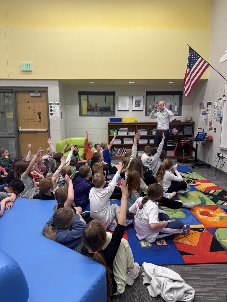A male guest speaker stands at the front of a classroom, gesturing with his hands as he addresses a large group of elementary students. The students are sitting on a colorful, patterned rug, and several have their hands raised enthusiastically to ask questions or participate.