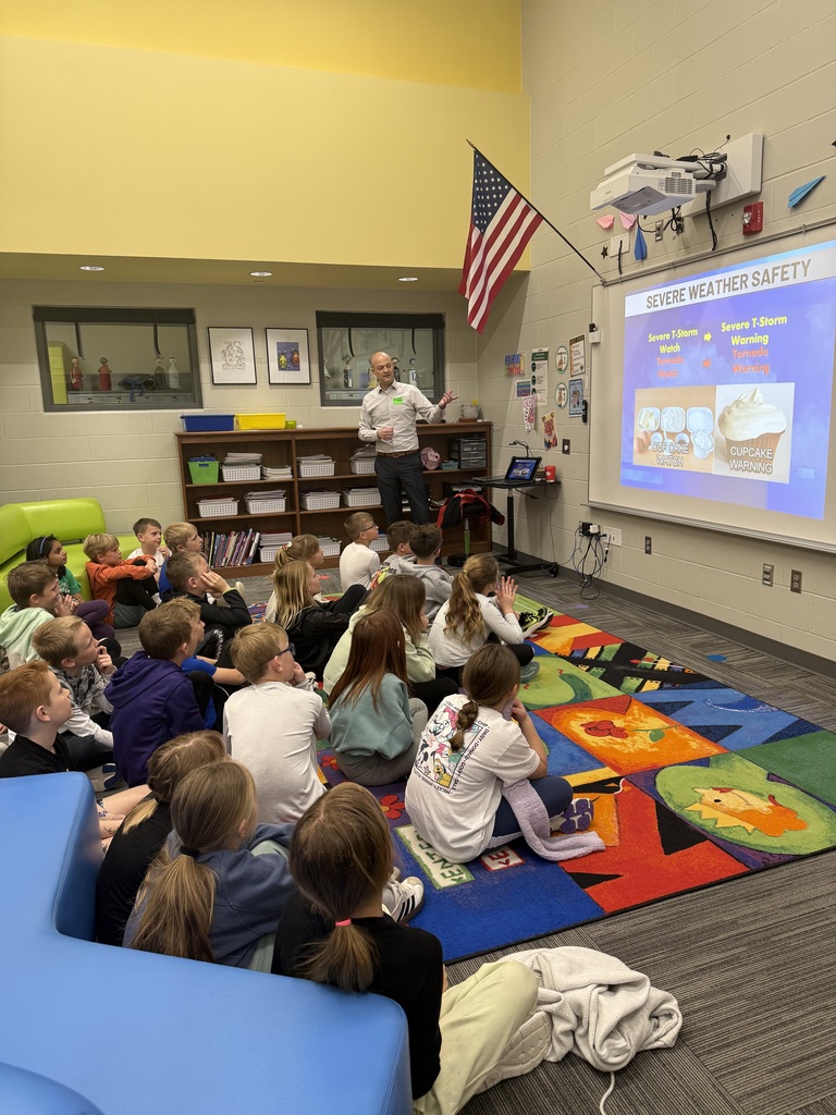 A classroom scene where a guest speaker points toward a projector screen titled "SEVERE WEATHER SAFETY." The presentation uses a "Cupcake Warning" analogy to explain weather alerts. A group of attentive students sits on a bright, multi-colored rug, looking toward the speaker and the screen.