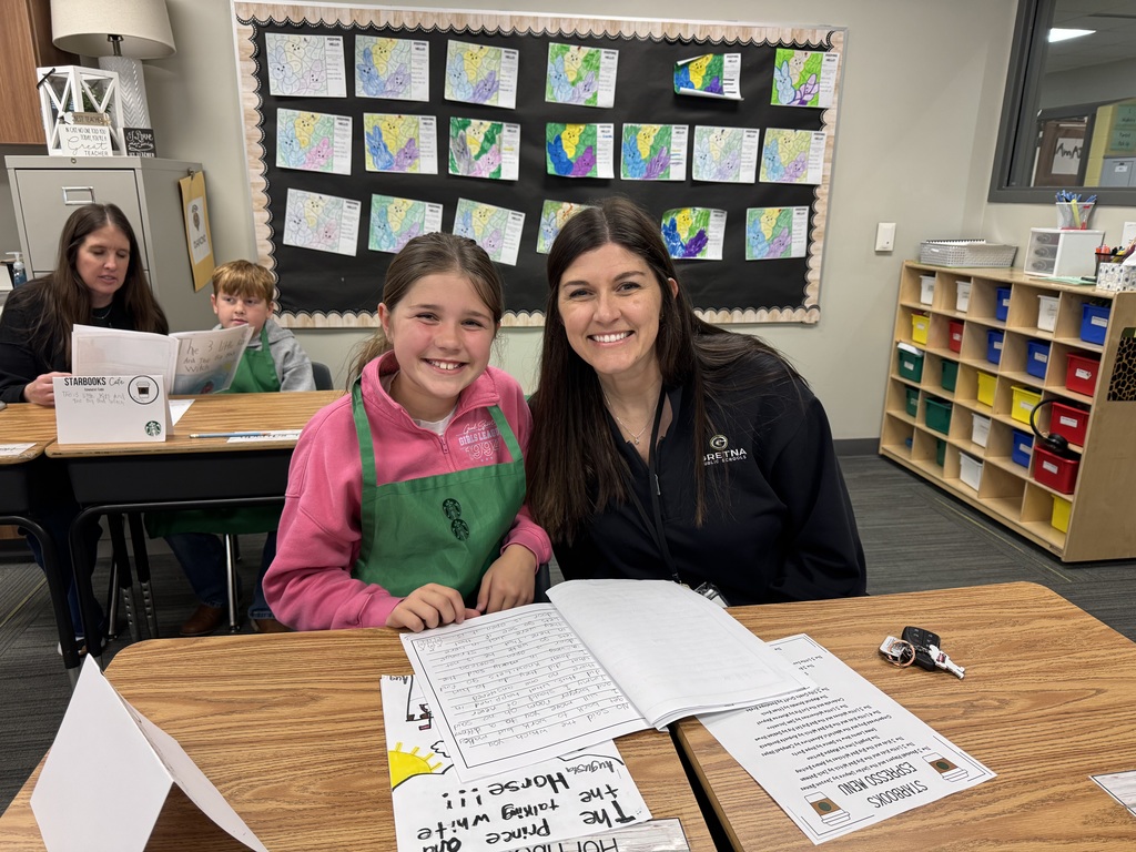 A smiling elementary student in a pink pullover and green apron sits at a desk with her teacher or a family member. They are looking at an open notebook filled with handwriting. A "Starbooks Espresso Menu" is visible on the desk.