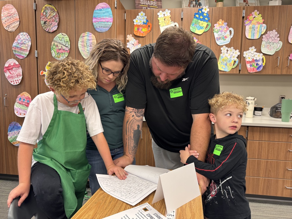 A student in a green apron and two adults lean in close to read a handwritten story in a notebook. A younger sibling stands nearby, also wearing a visitor sticker. The background is decorated with colorful paper Easter eggs and coffee cups.