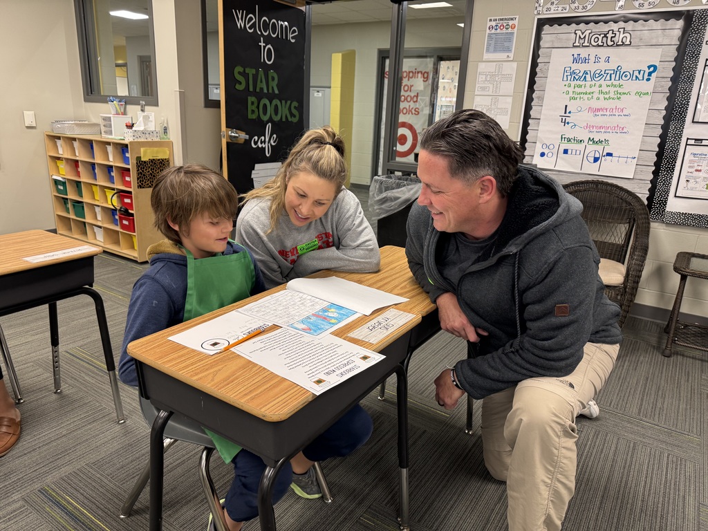 A young boy in a green apron sits at his desk, proudly showing his illustrated writing to two adults. The man, kneeling beside the desk, and the woman are both smiling warmly at the student’s work. In the background, a door is decorated with a sign that reads "Welcome to STAR BOOKS cafe."