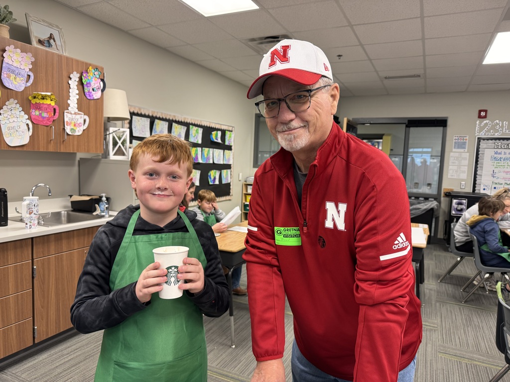 A student wearing a green apron holds a Starbucks cup and smiles at the camera, sporting a faint "milk mustache." He is standing next to a man wearing a red Nebraska pullover and a white "N" baseball cap.