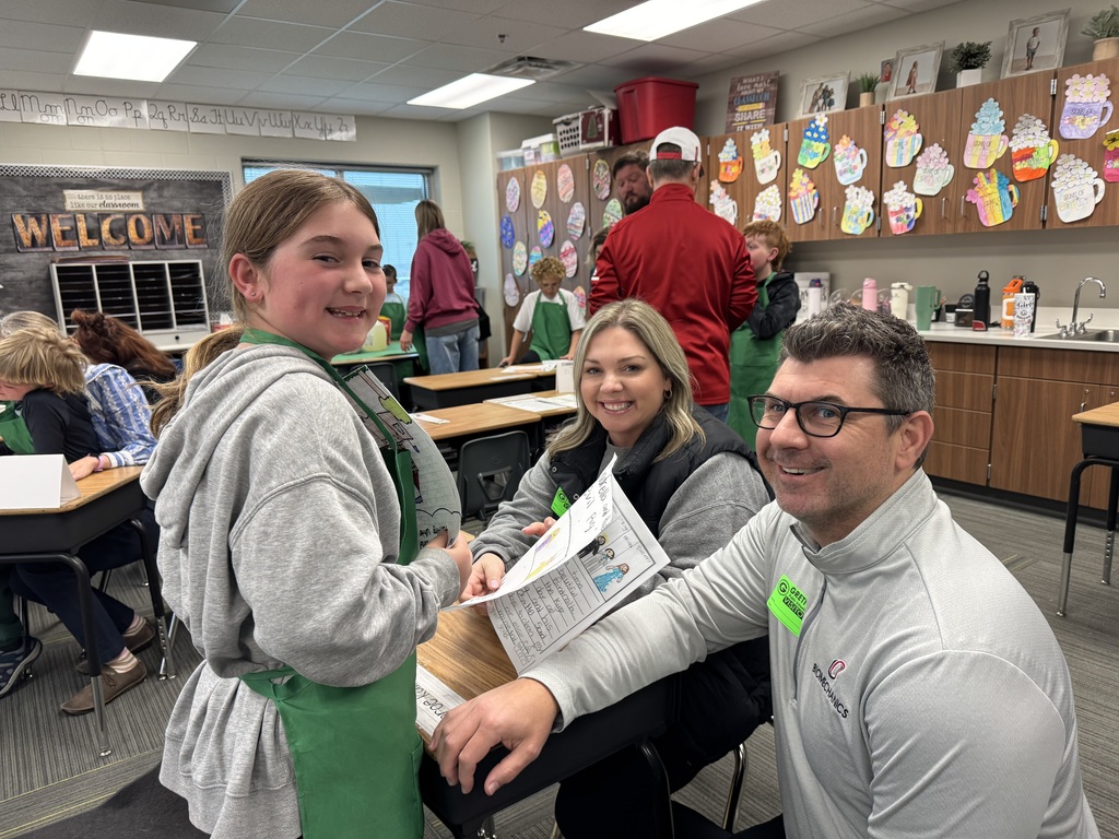 A young girl in a green apron stands at a desk, smiling as she presents her writing to two seated adults. The classroom background features coffee-cup-themed decorations on the cabinets and other families interacting.