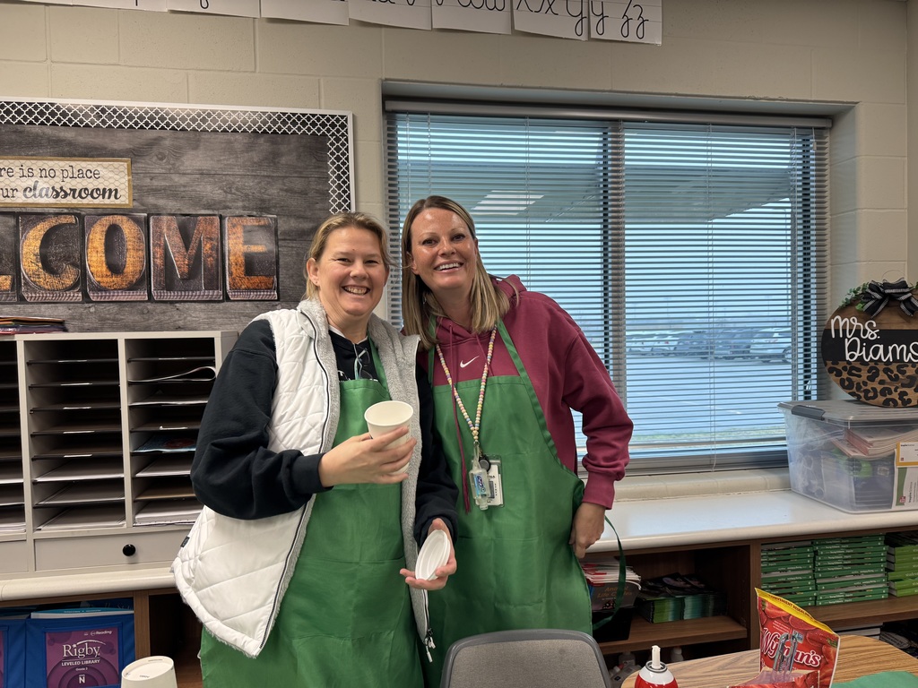 Two female teachers stand in a classroom smiling and wearing green "Starbooks" aprons. One holds a white coffee cup and lid, standing in front of a "Welcome to our Classroom" sign.