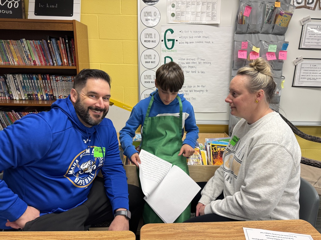 A student in a green apron leans over a notebook, pointing to his work as he reads it to two adults. The man on the left wears a bright blue Nebraska baseball hoodie and a "Gretna Visitor" sticker, smiling proudly.