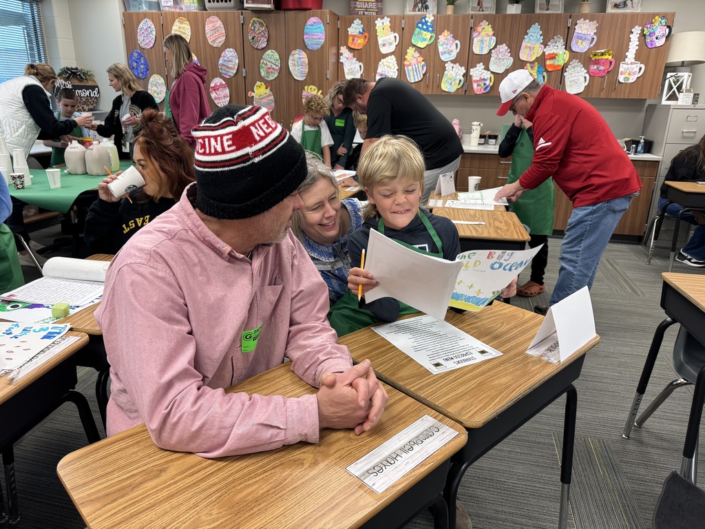 A student holding up a drawing of "The Big Blue Ocean" shares his story with two adults at a classroom desk. The classroom is buzzing with activity in the background, featuring cabinets decorated with colorful paper coffee mugs and other families enjoying the publishing party.