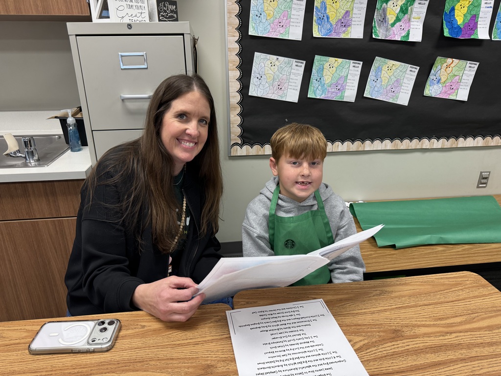 A young boy wearing a green apron sits at a desk next to a woman. They are both smiling at the camera while holding up a packet of the student's writing.