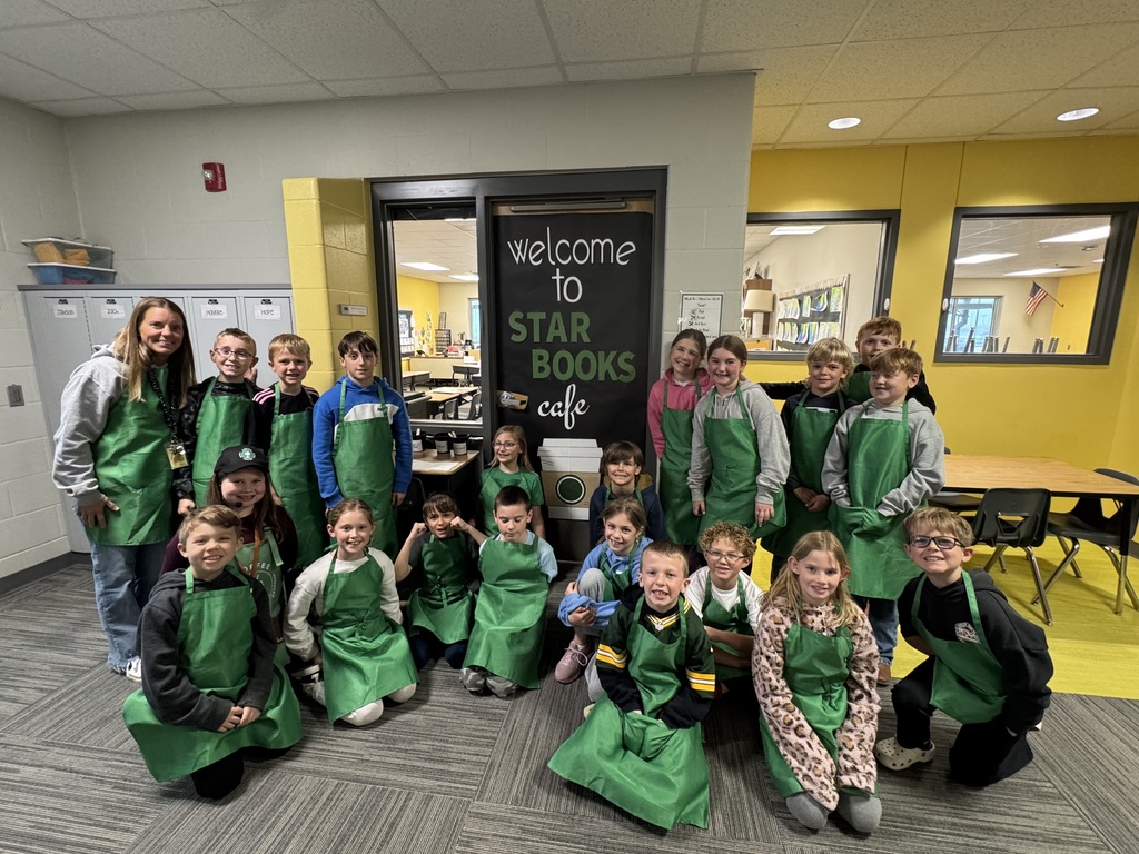 A large group photo of an elementary class and their teacher posing in the hallway. Almost every student is wearing a green "barista" style apron. They are gathered in front of a classroom door featuring a large black paper sign that says "Welcome to STAR BOOKS cafe" with a Starbucks-style coffee cup illustration.