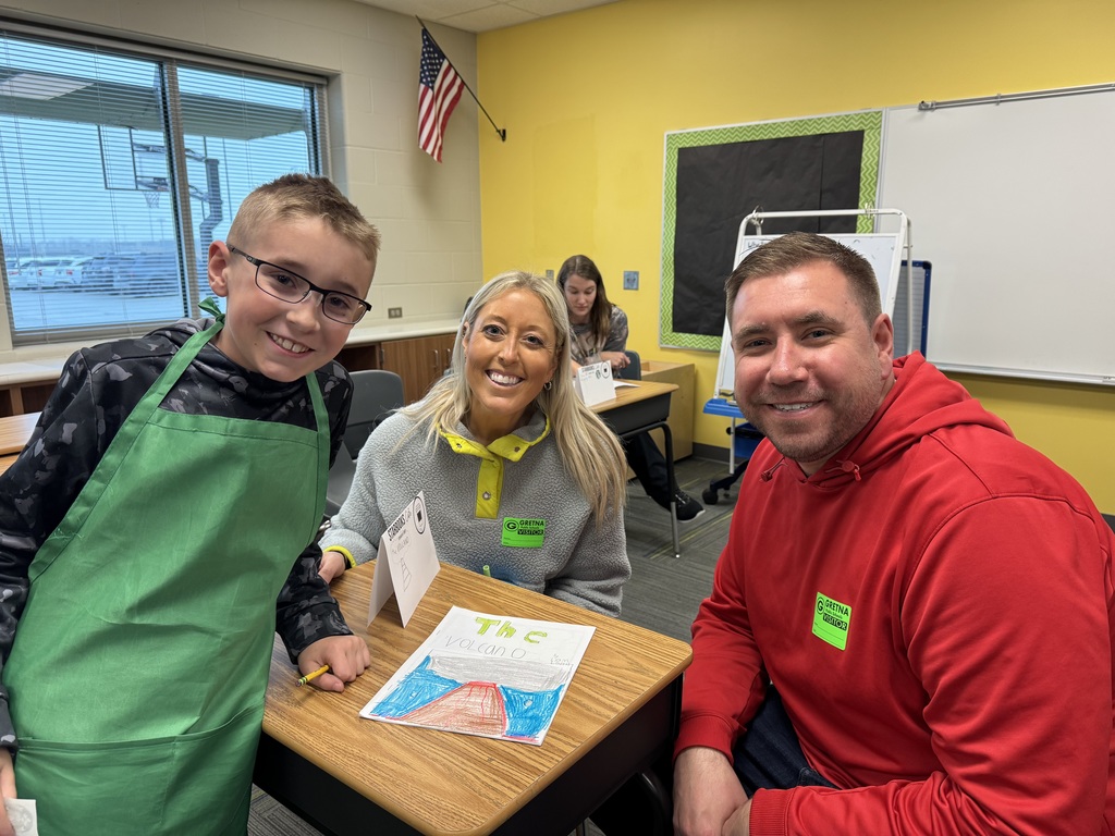 A student wearing a green apron points to a specific sentence in his notebook while reading aloud to a woman sitting beside him at a classroom desk.