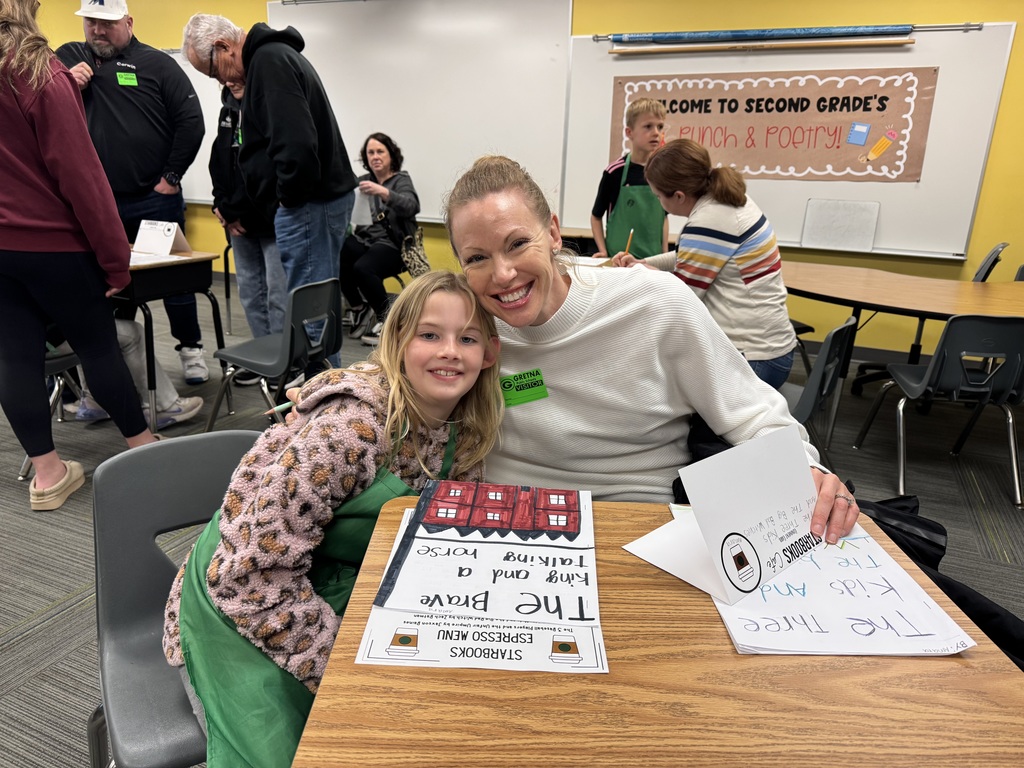 A smiling student stands beside a seated woman at a classroom desk. The desk is decorated with a "Starbooks Espresso Menu" and several handwritten stories with colorful illustrations.