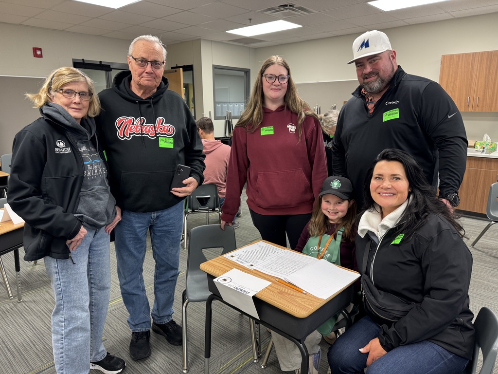 A student in a green apron and an adult male guest lean over a desk together, intently reading through a stapled packet of the student's writing.