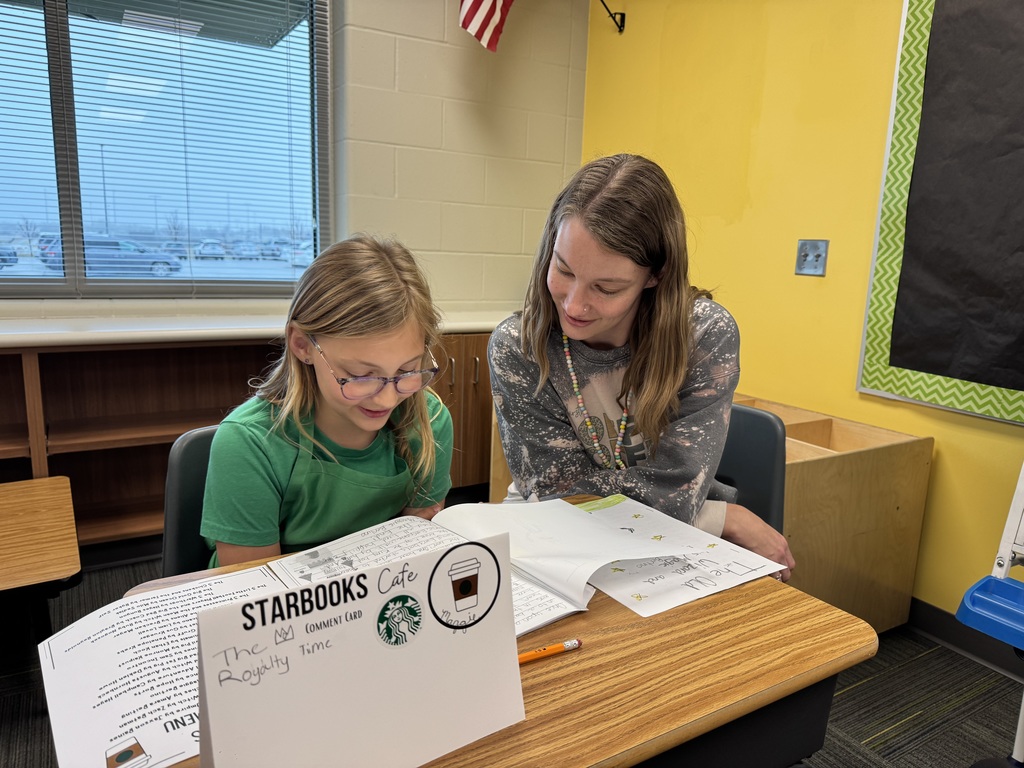 A young girl in a pink shirt and green apron sits at a desk with two adults, smiling happily for a photo. A "Starbooks Espresso Menu" and her writing assignments are laid out in front of them.
