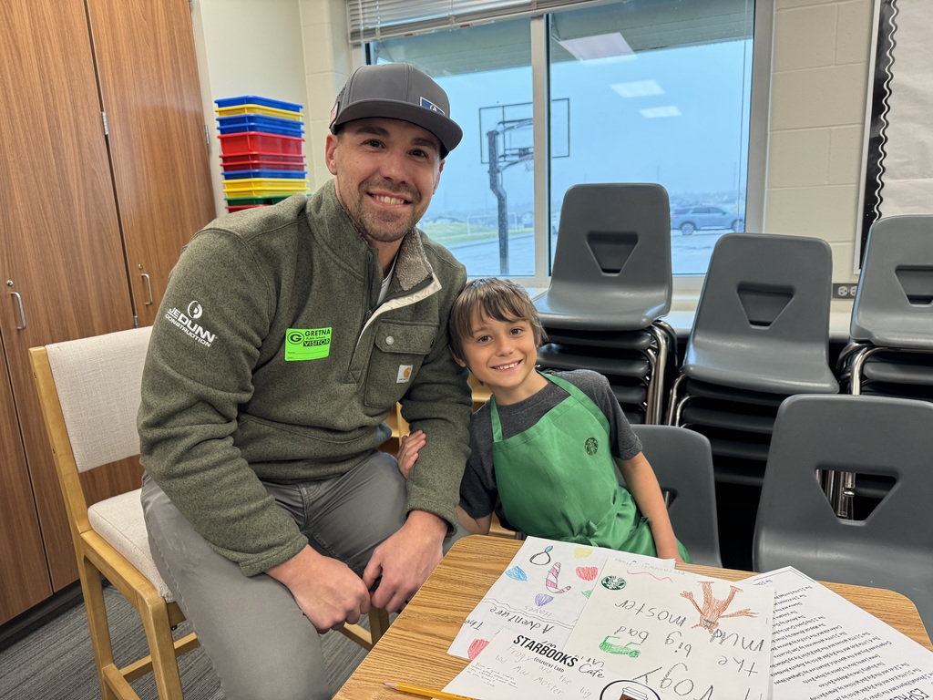A close-up of a student in a green apron sitting with two adults. They are all looking at the student's open notebook, which contains a story about a "Little Dog."
