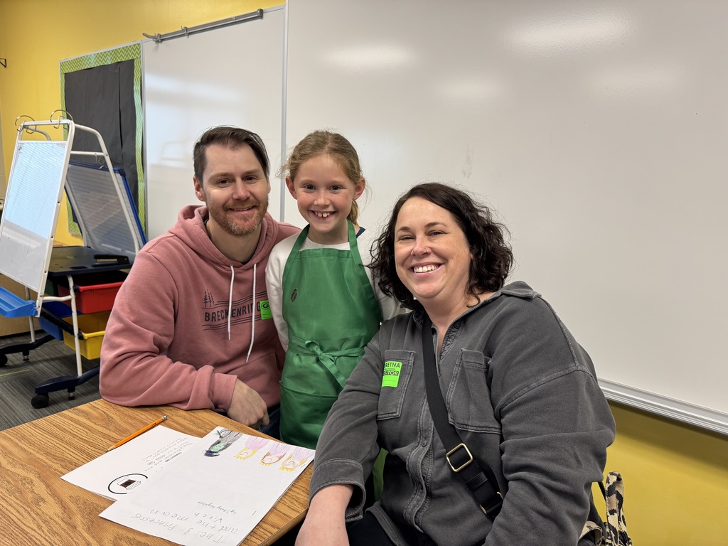 A student wearing a green apron and a woman sit together at a desk, looking at the camera and smiling. The student is holding a white coffee cup, and his writing packet is visible on the desk.