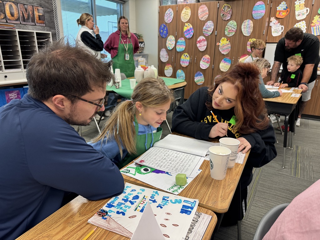 A smiling student stands beside a seated woman at a classroom desk. The desk is decorated with a "Starbooks Espresso Menu" and several handwritten stories with colorful illustrations.