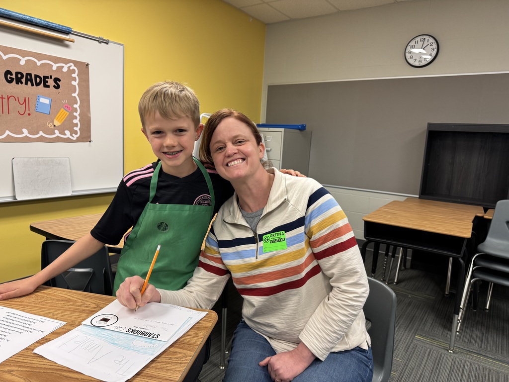 A student in a green apron smiles while sitting at a desk with an adult. They are holding a Starbucks-branded paper cup and a packet of the student's writing titled "The Three Little Pigs."