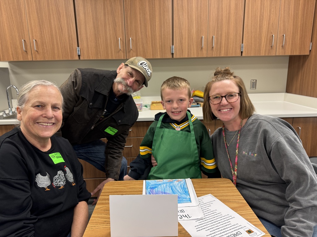 A young boy wearing a green apron smiles brightly as he sits between two adults. They are gathered around his desk, which displays his "Starbooks" writing projects and a "Gretna Visitor" sticker on the man's shirt.
