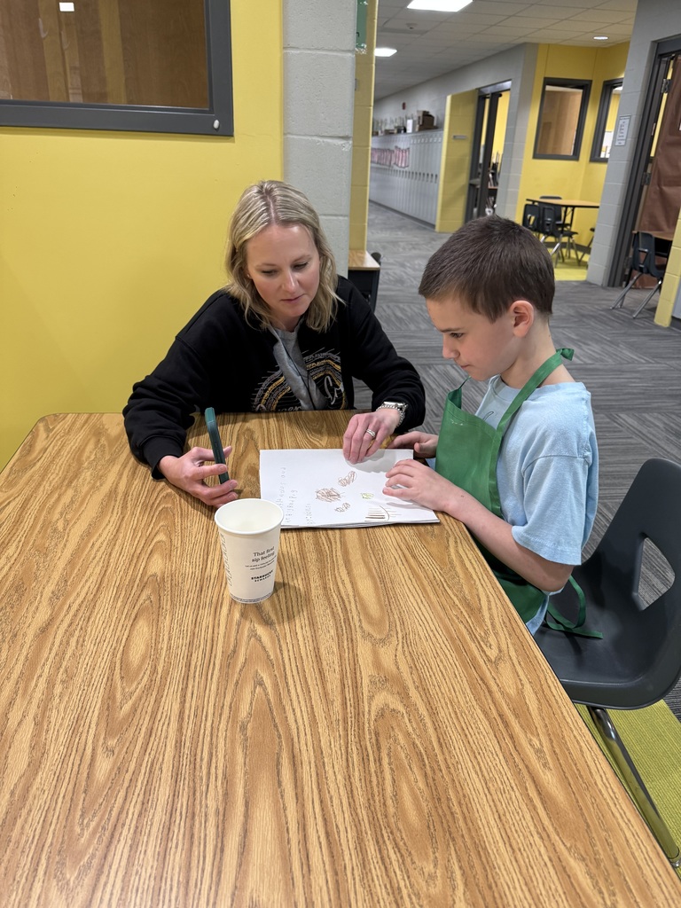 An elementary student wearing a green apron sits at a desk and looks up with a smile while sharing a notebook of handwritten work with a woman sitting next to him.