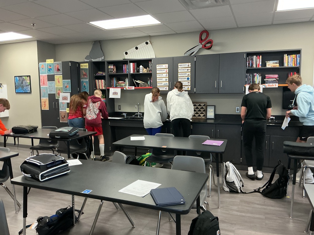 Photo of students moving around the classroom identify which Greek gods go with which cabin photos hanging on the walls.