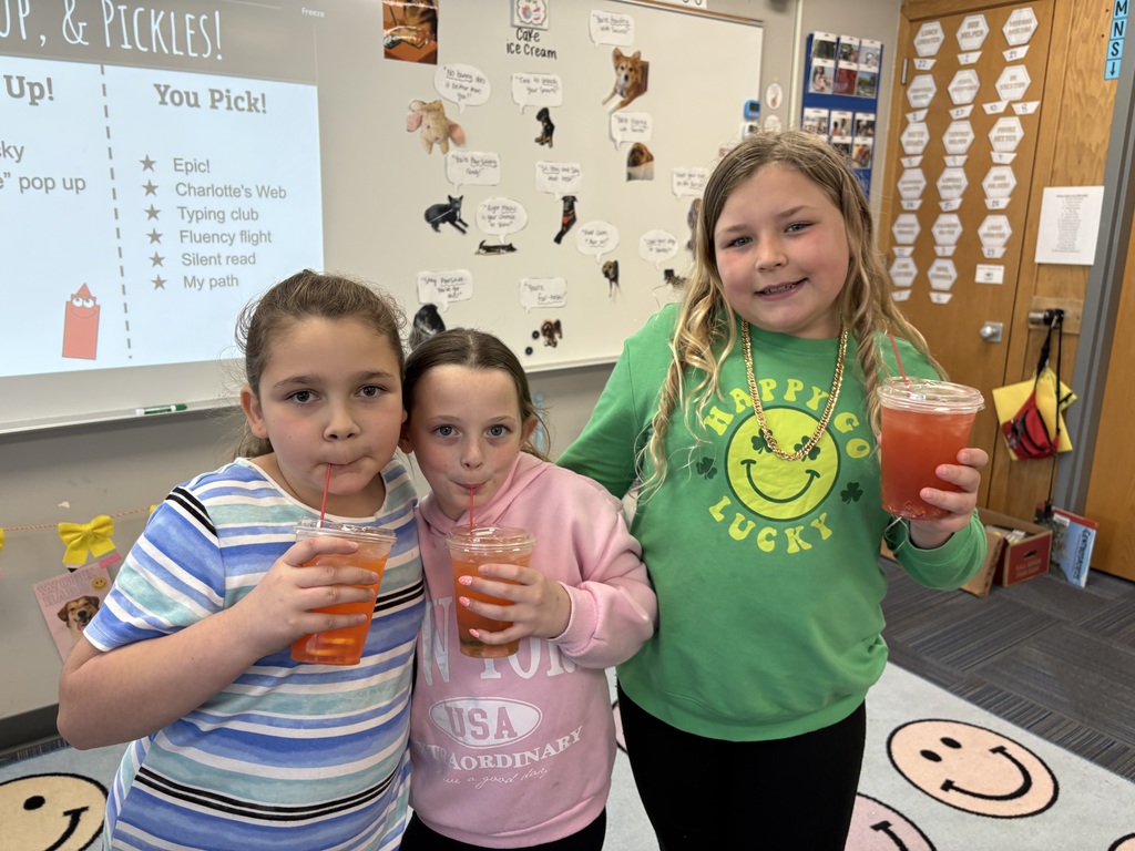 Three girls standing together in a classroom; two are sipping red drinks through straws while the third, in a green "Happy Go Lucky" shirt, smiles.