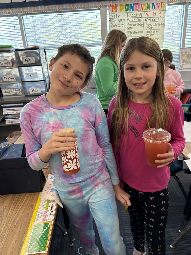Two young girls posing in a classroom; one wears a tie-dye set and holds a floral glass, while the other wears pink and holds a plastic cup of red juice.