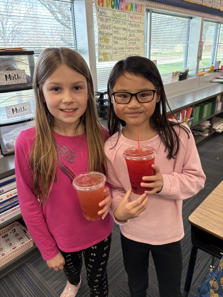 Two girls standing side-by-side in a classroom holding clear cups of red juice; one is in a bright pink shirt and the other is in a light pink sweater with glasses.