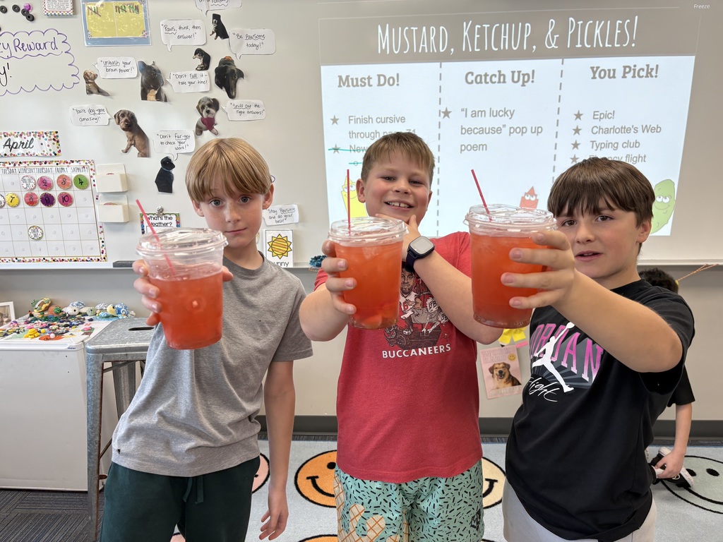 Three boys standing on a smiley-face rug, smiling and holding up clear plastic cups filled with red iced drinks toward the camera.