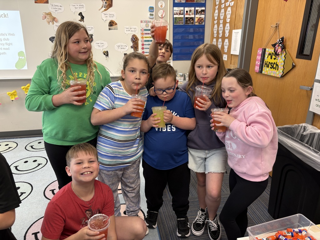A group of six children huddled together in a classroom, smiling and posing while enjoying their colorful iced drinks.