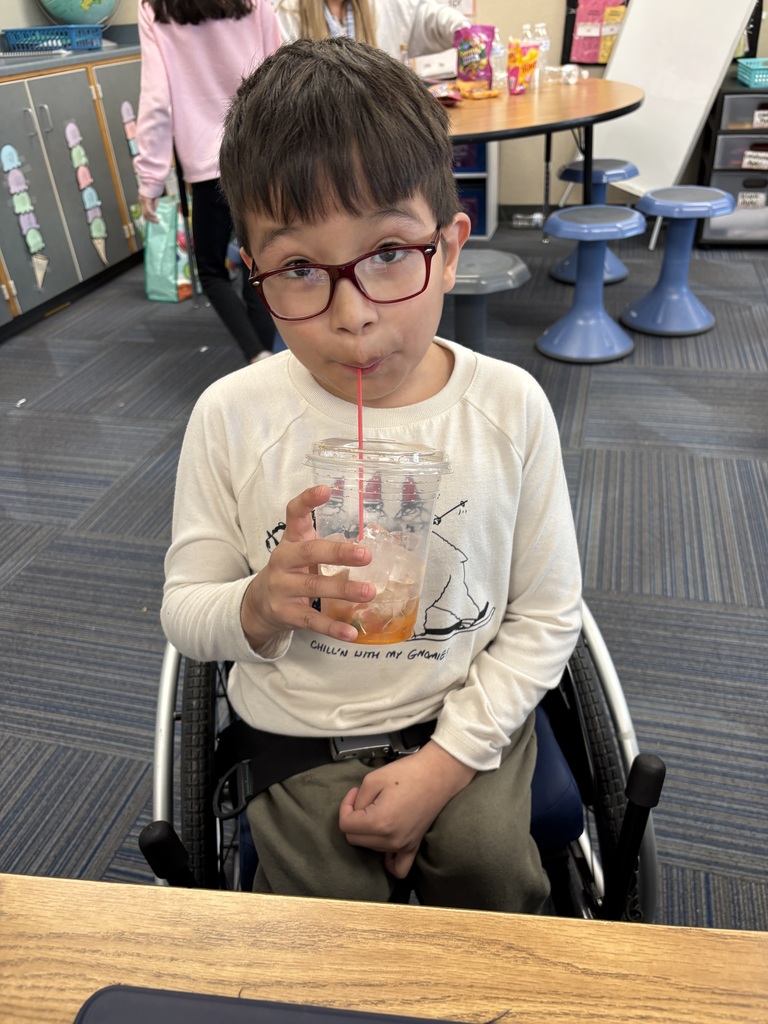 A young boy in a wheelchair wearing glasses and a white long-sleeve shirt, sipping a red iced drink through a straw.