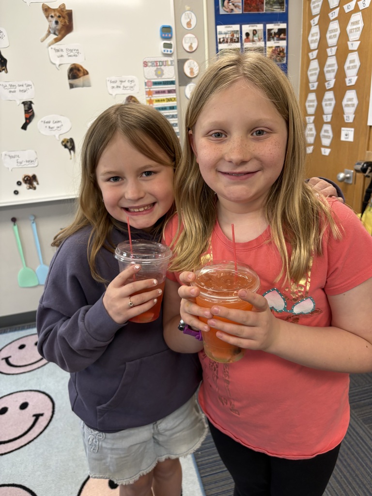 Two young girls with blonde hair standing close together and smiling, both holding clear plastic cups of orange-red juice.