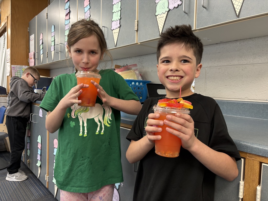 A girl in a green unicorn shirt and a boy in a black t-shirt posing with their drinks; the boy's cup is topped with gummy worm candies.