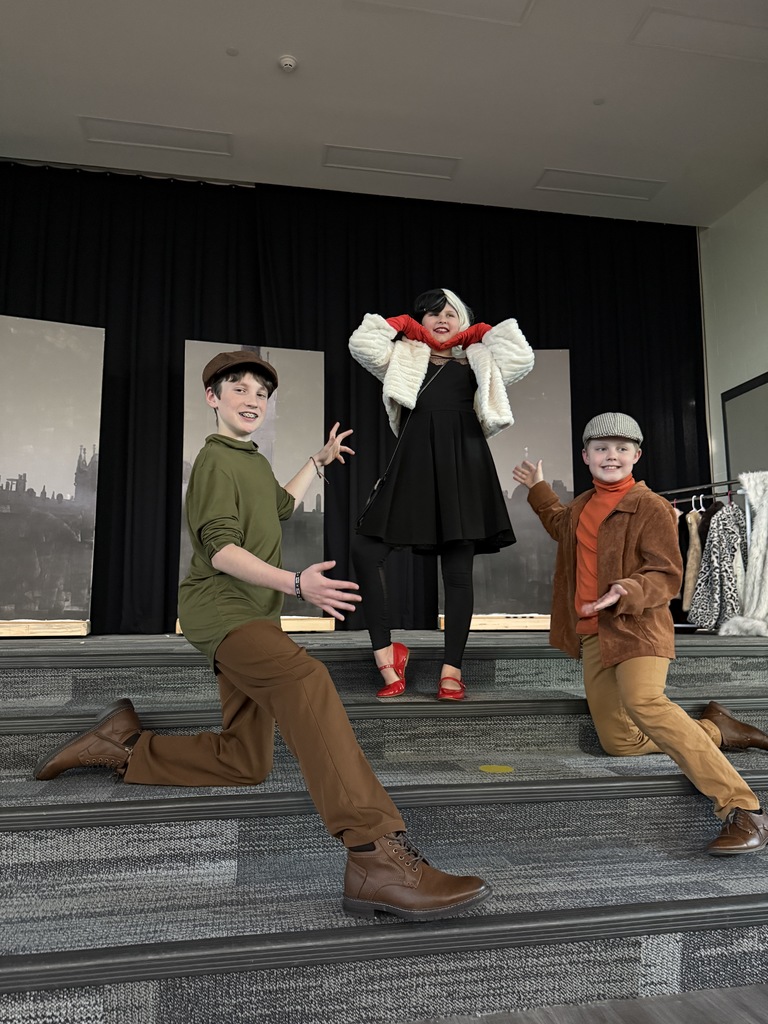 A boy in a green shirt and brown pants is kneeling with hands out, a boy with brown pants, orange jacket and hat is kneeling and a girl dressed as Cruella DeVille is in the middle.