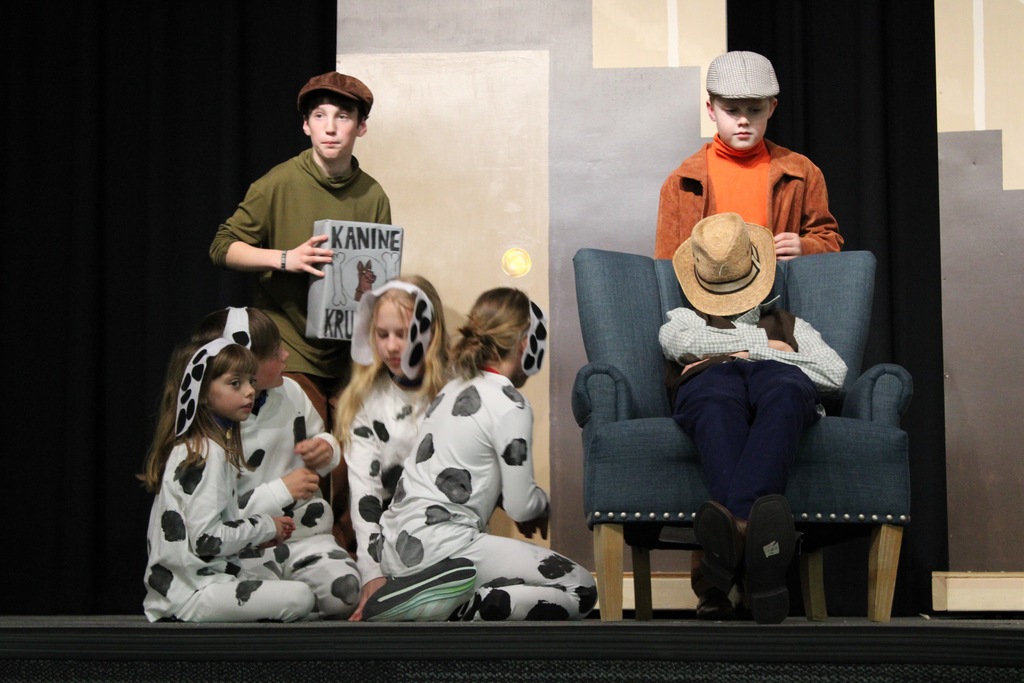 A boy with a green shirt and brown hat is holding a gray kanine krunchies box with 4 kids dressed as dalmatians in front of him. A farmer sleeps in a blue chair next to the dogs with a boy in an orange shirt, brown jacket and gray hat behind.